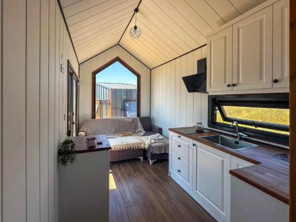 Tiny house salon ve mutfak alanı; büyük pencere, beyaz dolaplar ve ahşap tezgah.